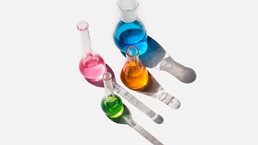 Laboratory glassware containing liquids of different colors on a light gray background. 