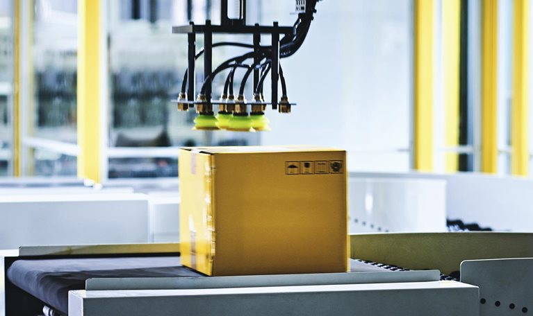 Close-up of robotic arm carrying cardboard box on conveyor belt at modern distribution warehouse.