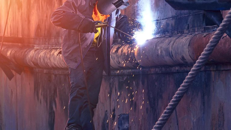 Welder in protective workwear on scaffold is welding the old rusty vessel hull in shipyard area at sunset time