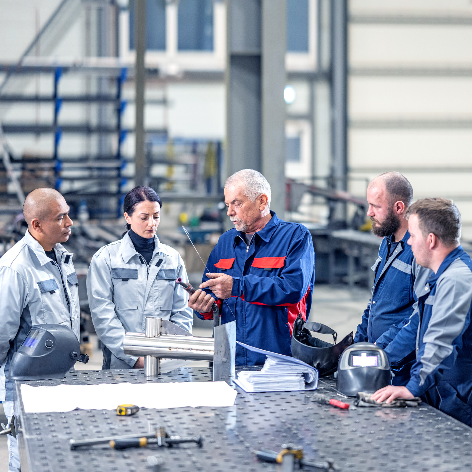 A group of four employees wearing blue and grey coveralls are gathered around a senior colleague in a metal workshop. The senior staff member is holding a piece of part and gesturing towards it as they explain something to the others.