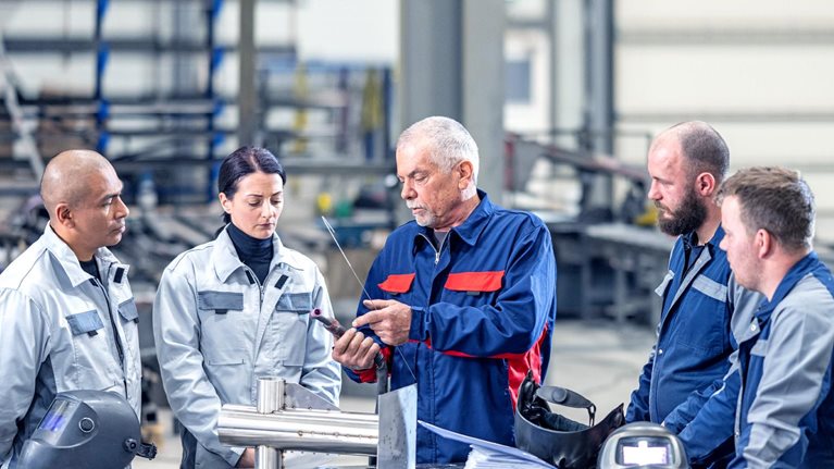 A group of four employees wearing blue and grey coveralls are gathered around a senior colleague in a metal workshop. The senior staff member is holding a piece of part and gesturing towards it as they explain something to the others.