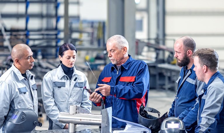 A group of four employees wearing blue and grey coveralls are gathered around a senior colleague in a metal workshop. The senior staff member is holding a piece of part and gesturing towards it as they explain something to the others.