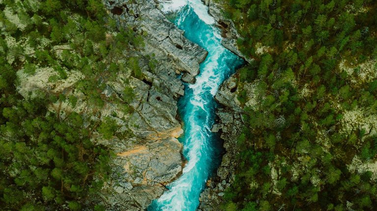 Drone high-angle photo of the turquoise-colored mountain river flowing in the pine woodland with a view of the mountain peaks in the background in Innlandet County, Norway