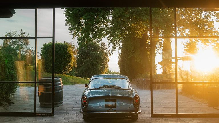 Rear view of classic car driving out from behind glass garage doors as sun sets in background.