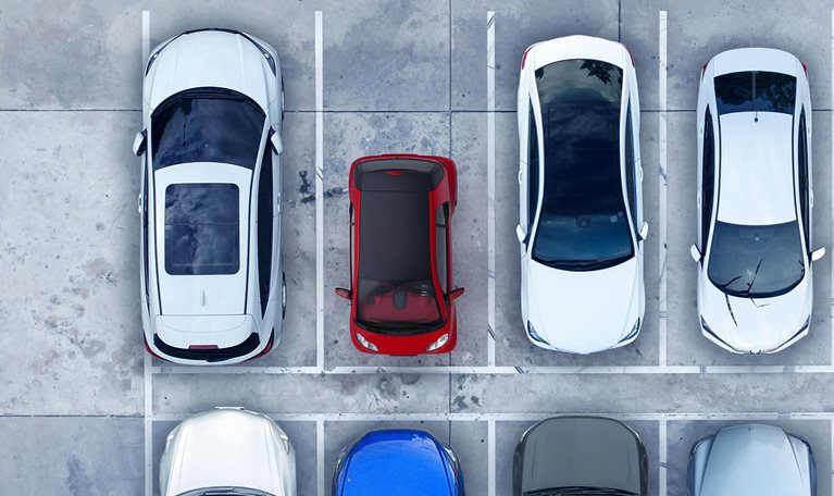 An overhead view of a red, ultra-light microcar parked among 7 full size vehicles highlighting the contrasts in size and space between them