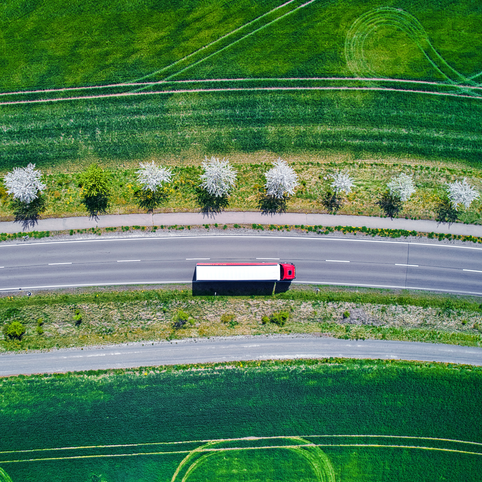 Aerial view of a red and white semi-truck driving down an interstate road though green farm fields