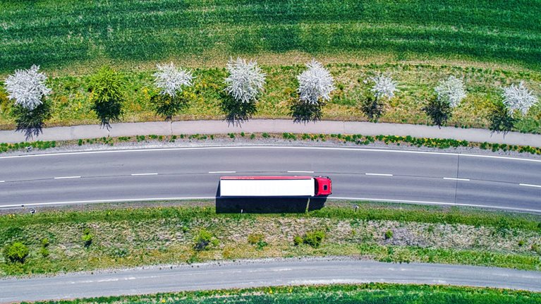 Aerial view of a red and white semi-truck driving down an interstate road though green farm fields