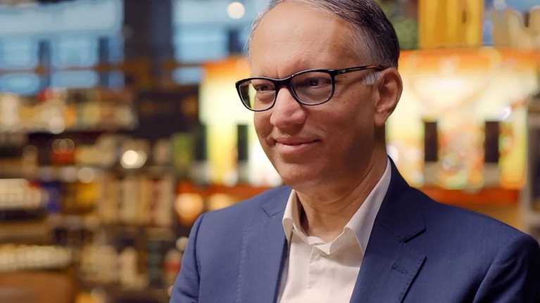 Vipul Chawla in a blue suit and glasses sits in a warmly lit store or café setting, smiling slightly, with shelves and lights blurred behind.