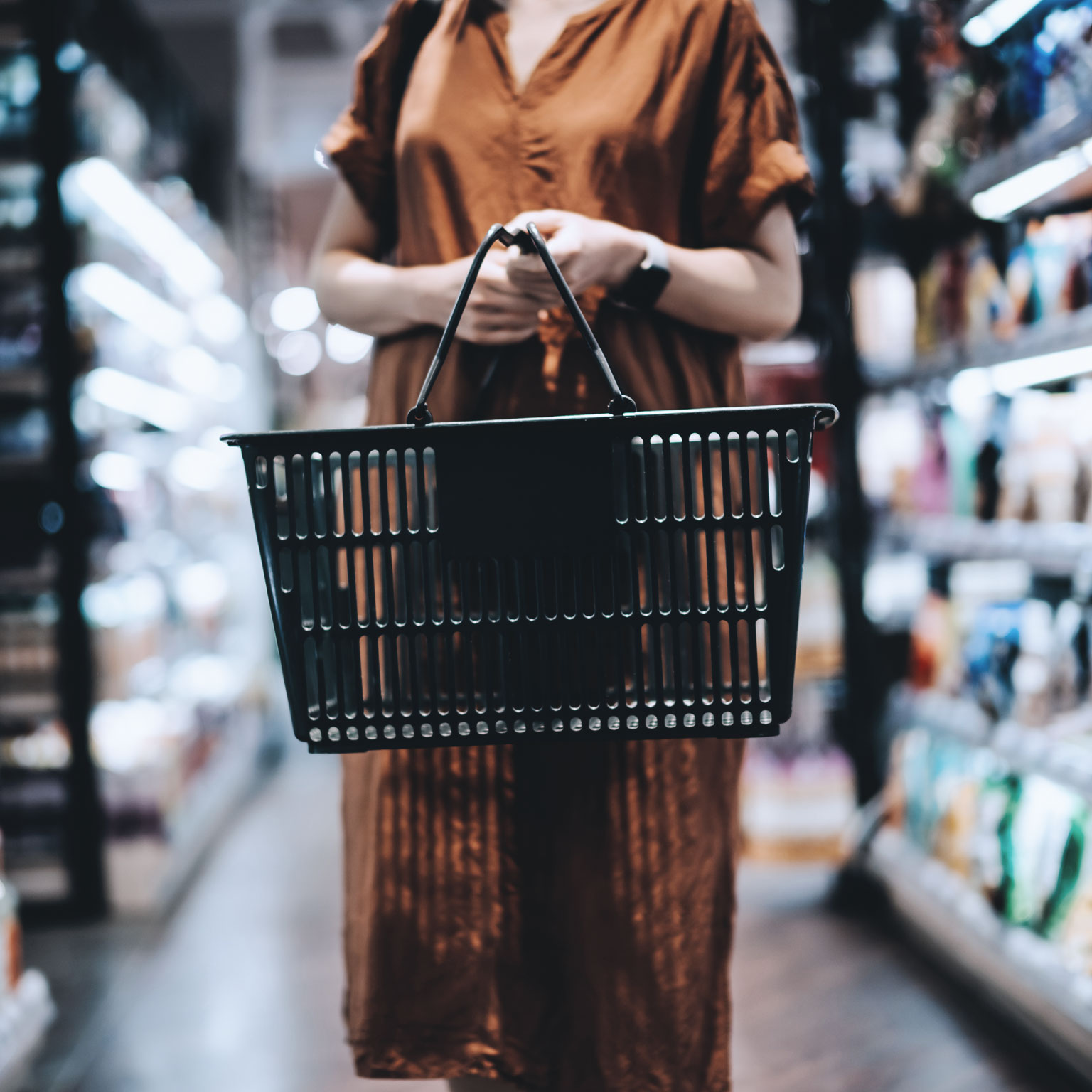Woman standing in a grocery store aisle holding a black shopping basket. 