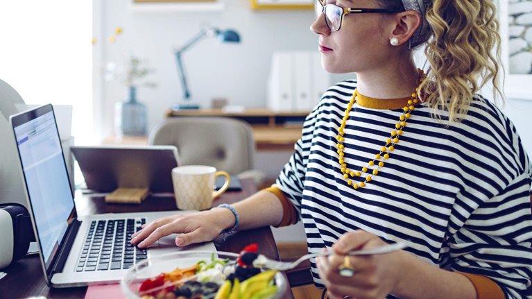 A woman wearing glasses and a striped shirt is seated at a desk, looking at her laptop and eating a salad.