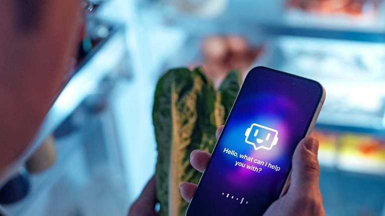 A person standing in front of an open refrigerator while holding a smartphone in one hand and a head of romaine lettuce in the other. The phone screen displays a glowing chatbot interface with the message, “Hello, what can I help you with?”.