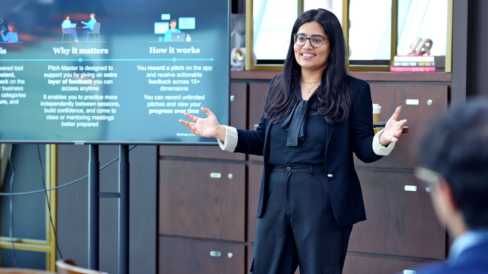 A person with long dark hair and glasses in a dark business suit gestures with open arms while addressing an audience; behind them a large screen shows a presentation with headings “Why it matters” and “How it works.”