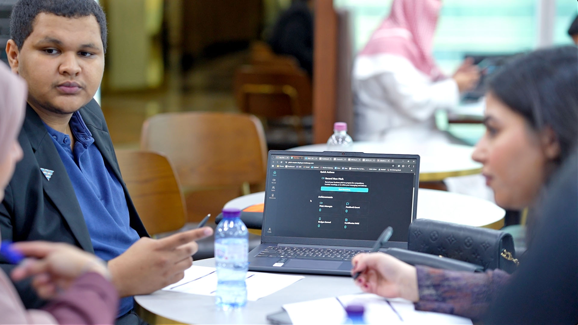 A person in a blazer holds a smartphone at a round table with a laptop displaying a user interface and a water bottle, while talking with another person; a third person wearing a headscarf is in the background.