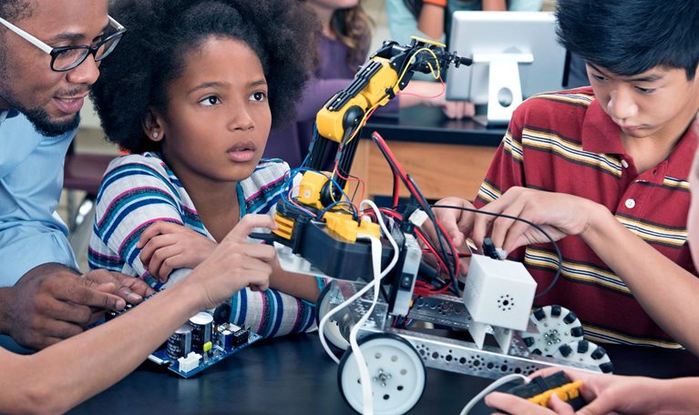 Children from diverse racial identities, who appear to be in middle school, are creating a mechanized forklift type of machine on wheels in a mechanics class with a teacher helping them.
