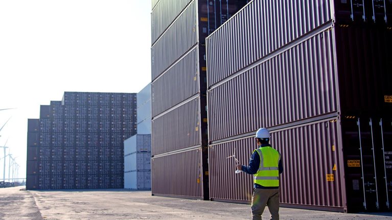 A worker in a dockyard, donning a helmet and reflective vest, gazes at a blueprint of a windmill. In the distance, there are rows of trucks and wind turbines.