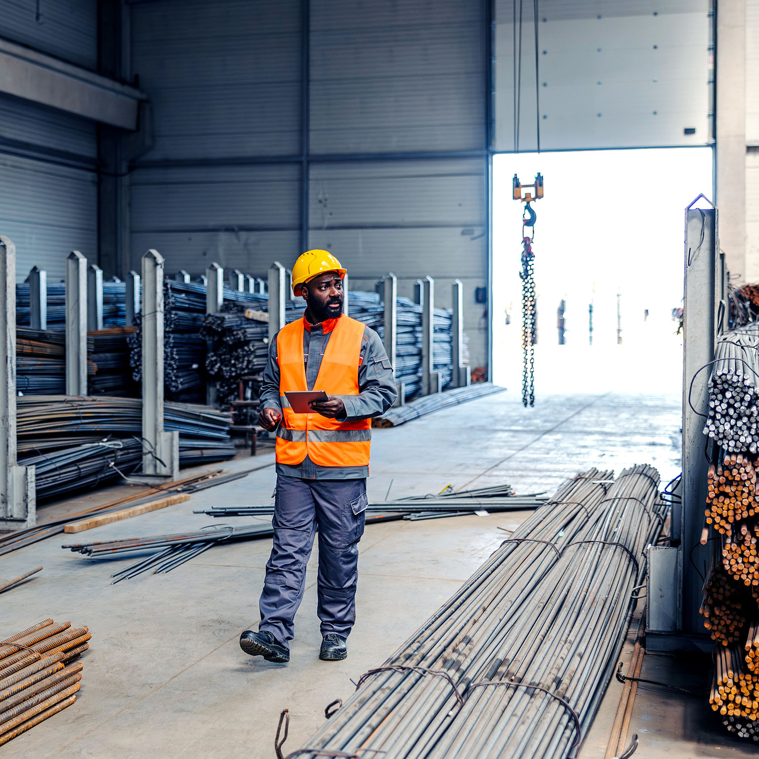 Man in a safety vest and hard hat walking through an industrial warehouse, holding a digital tablet and checking steel rebar inventory