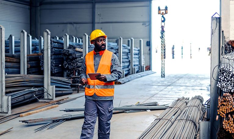 Man in a safety vest and hard hat walking through an industrial warehouse, holding a digital tablet and checking steel rebar inventory