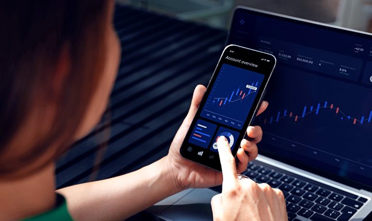 A person uses a smartphone displaying financial charts while viewing a matching investment dashboard on a laptop.