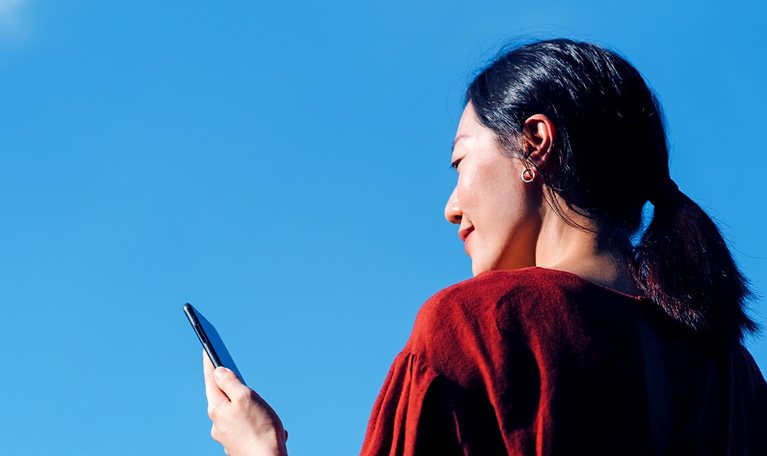 Low angle portrait of young Asian woman using smartphone against beautiful blue sky