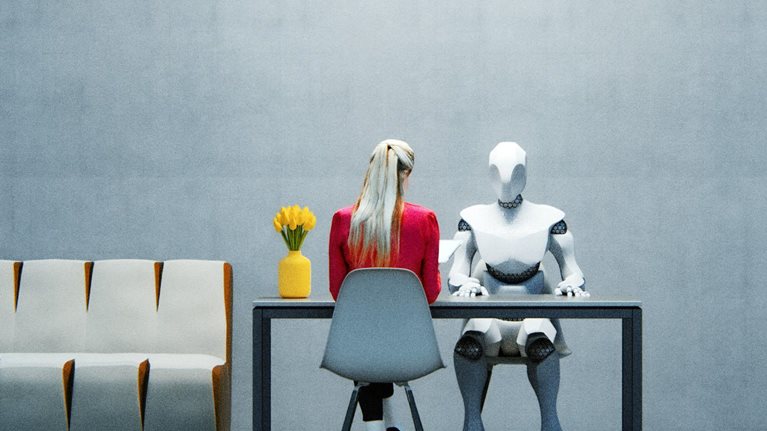 A woman in a red jacket sitting at a table across from a humanoid robot in a sparsely decorated office space.