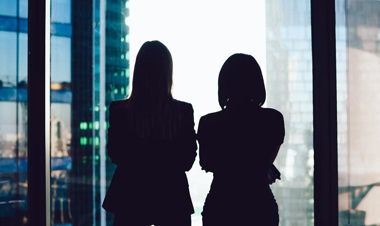 Back view of female colleagues in formal wear standing near window looking at modern exterior of skyscrapers in business center, silhouette of women together planning future success of brainstorming