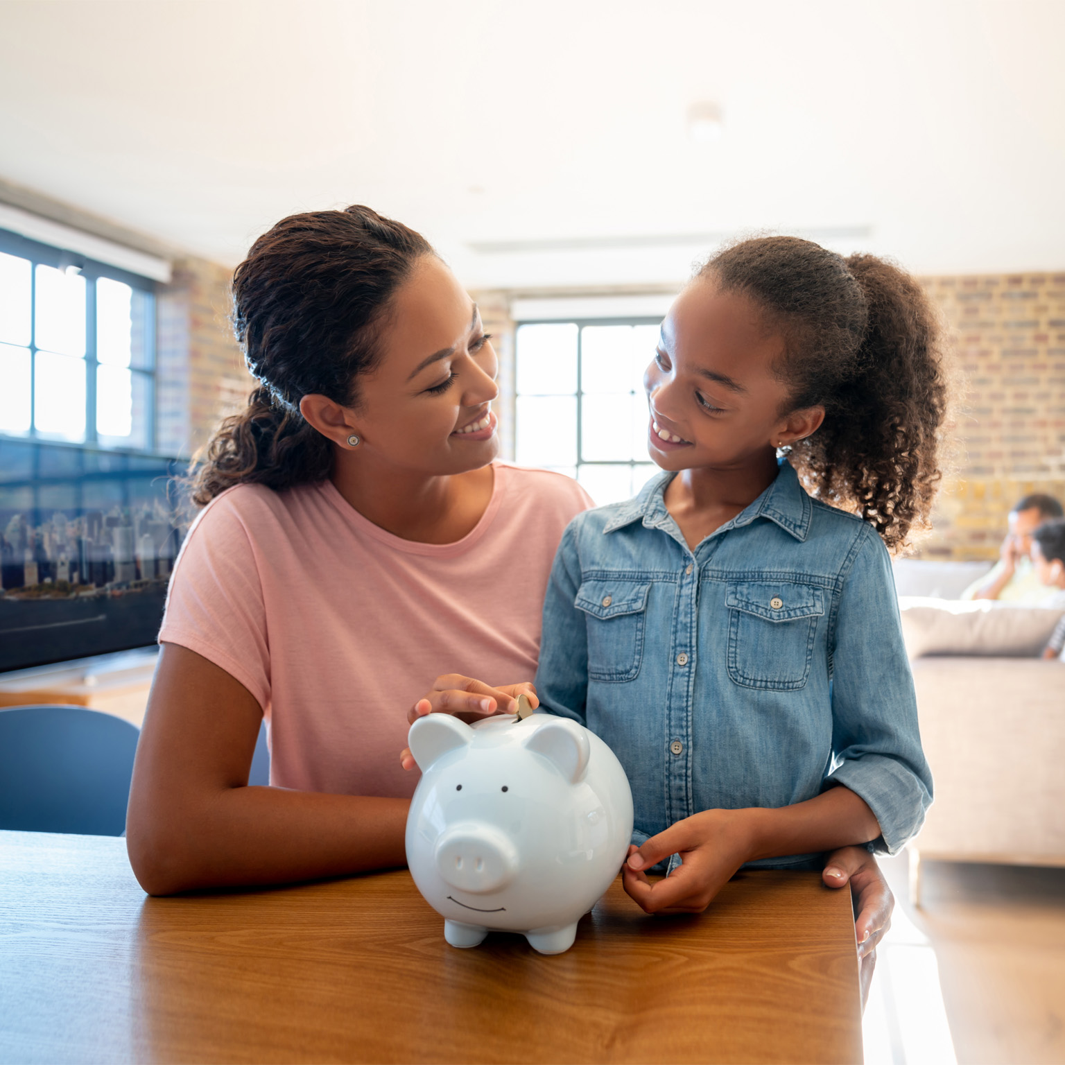 photo mother and daughter, both smiling, looking at each other as daughter drops coins into piggy bank
