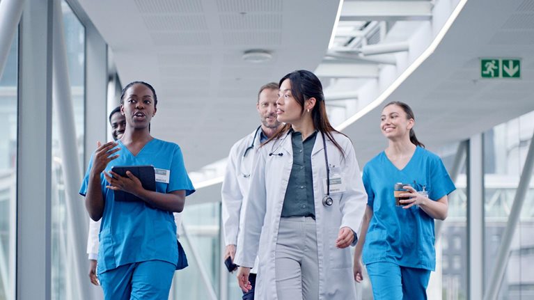 A doctor and nurse with tablet for discussion while leading a group of 5 peers that are walking down a hospital hallway together. It represents a diverse team of medical professionals utilizing technology in their collaboration.