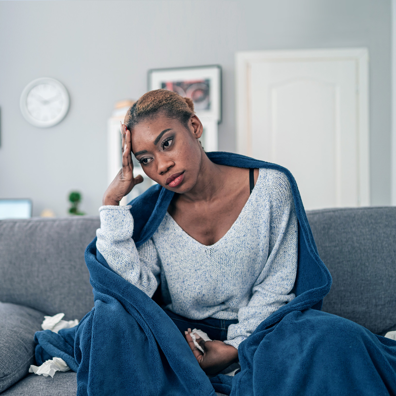 A young woman sitting on her couch wrapped in a blanket with used tissues laying next to her.