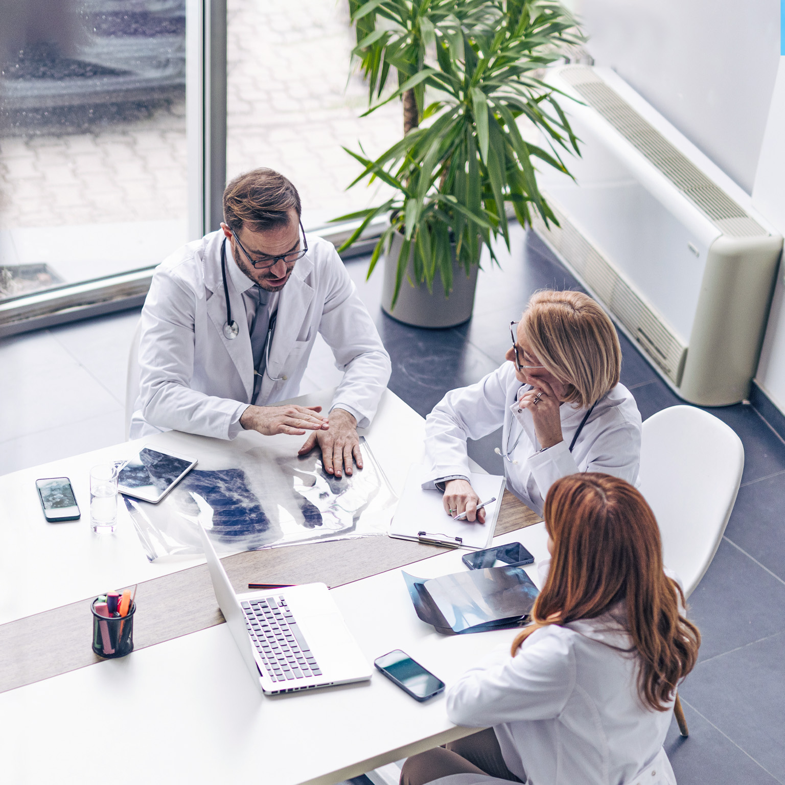 From a bird's-eye view, a diverse group of business professionals is gathered around a table covered with laptops, tablets, design plans, and financial reports. The team is engaged in a lively conversation.