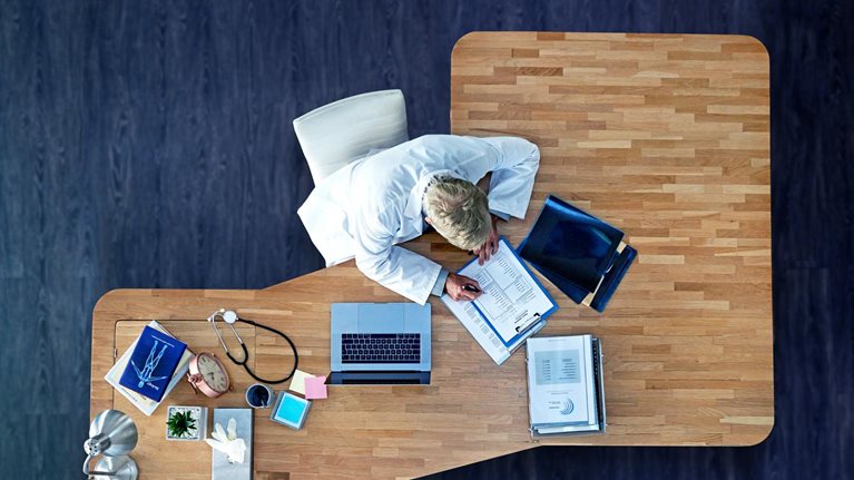 A bird's eye perspective of a physician sitting at an executive desk, filling out paperwork. Beside them sits a laptop, medical records, and a stethoscope. The desk is positioned on a rich, dark wooden floor.