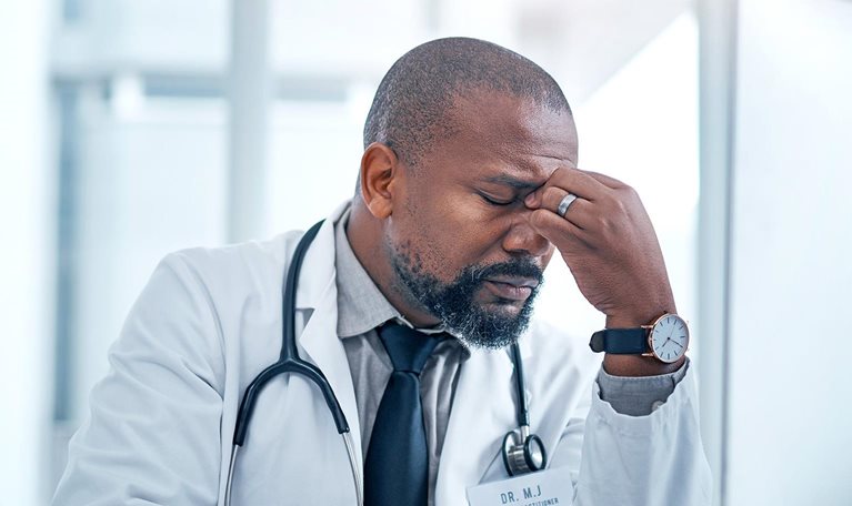 A Black male doctor sits at a computer looking frustrated