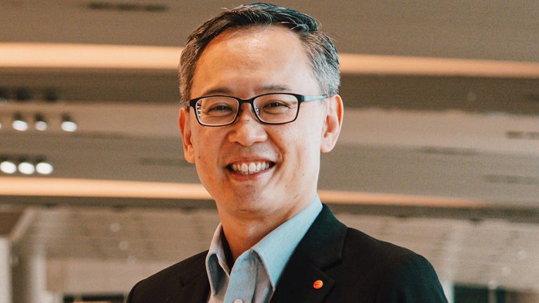 Lim Ching Kiat smiles while standing in an airport, dressed in a suit and wearing glasses.