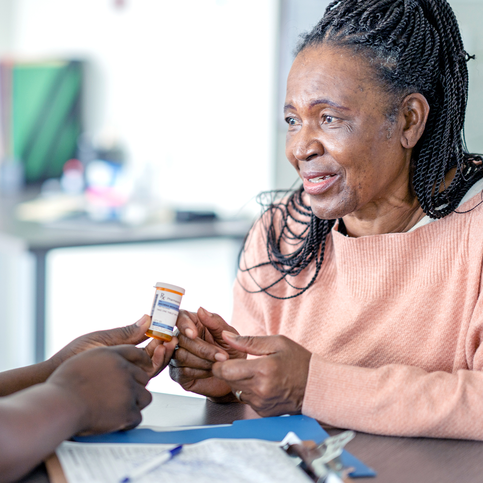 A senior woman sits across the desk from a nurse practitioner as they discuss her medications. She is dressed casually and looking at the nurse as she listens attentively. The nurse is dressed in blue scrubs and has a headscarf on as she holds out the medication and informs the woman about how to take the new medication.