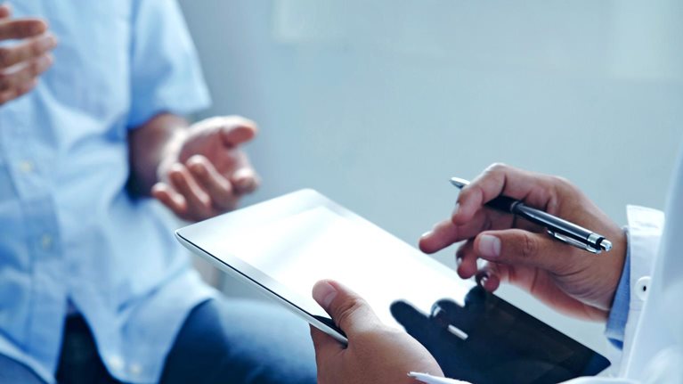 A close up of a doctor's hands holding a tablet while talking to a patient about his treatment plan.