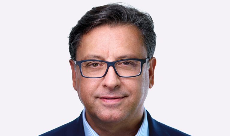 A man named Paul Hudson, sporting dark hair and glasses, can be seen in a slight smile as he faces the camera. He is dressed in a blue suit jacket and a striped blue shirt.