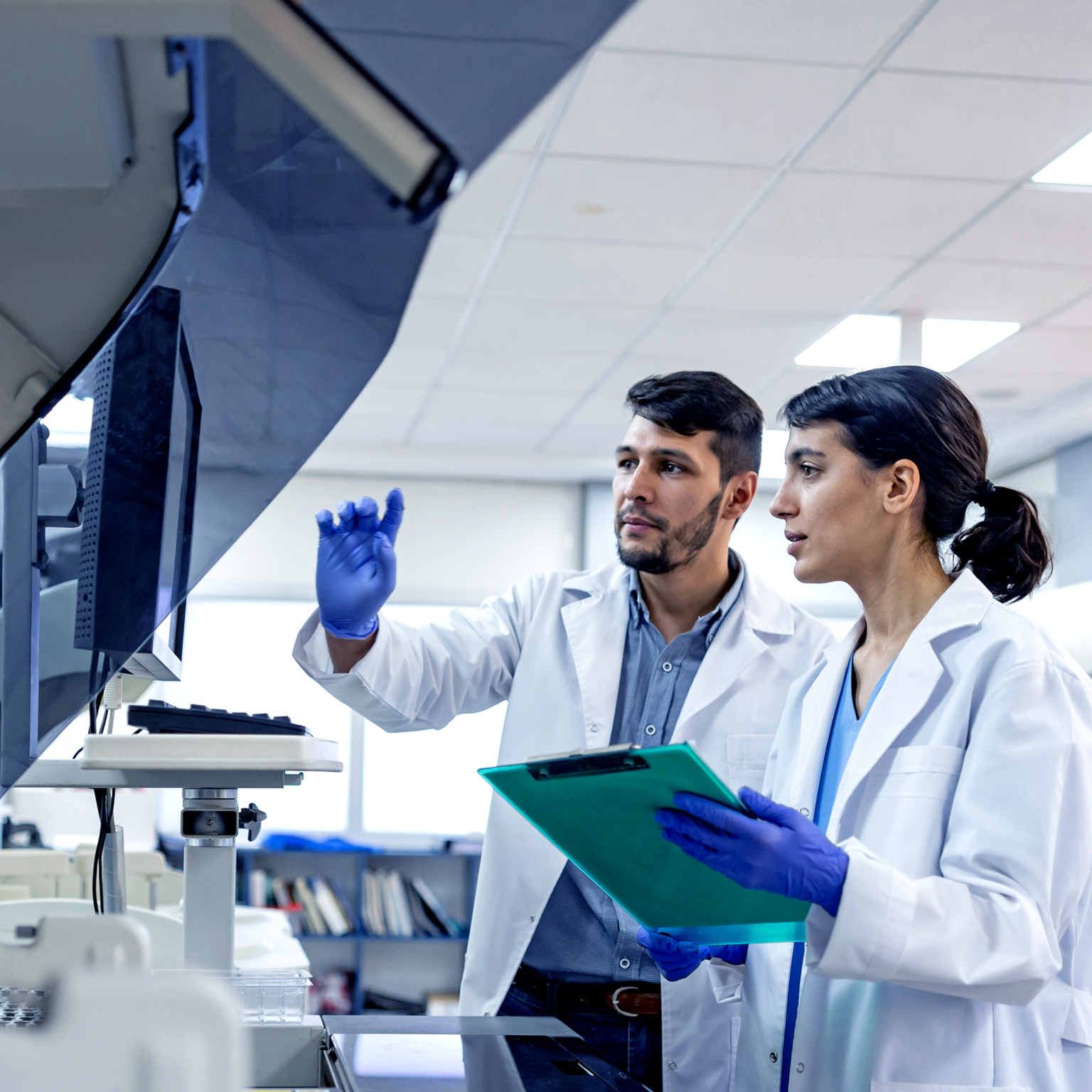 Two laboratory scientists in white coats and blue gloves stand side by side examining a large machine with a monitor. One scientist points to the screen while the other holds a clipboard, both appearing focused on the data or results displayed.