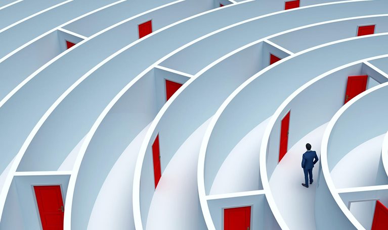 A man stands in the middle of a large maze that has many doors. A pathway out is not clear.