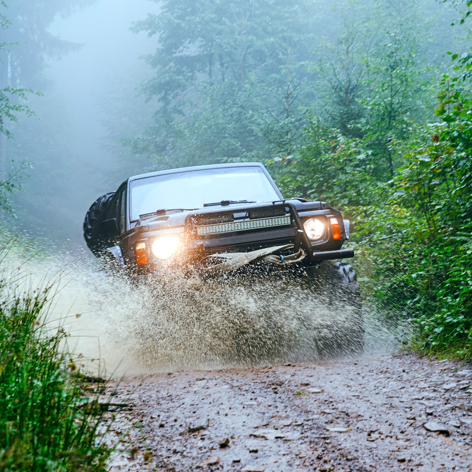 The image shows a rugged off-road vehicle driving through a muddy forest trail, splashing water as it moves forward. Dense green foliage surrounds the path, and mist hangs in the air, creating a dramatic and adventurous atmosphere.