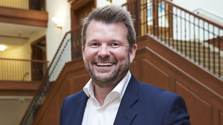 A portrait of Reynir Indahl, founder and managing partner of Summa Equity, in a dark blue suit standing in a grand, wood-paneled building with a large staircase in the background. He is leaning on a railing, looking directly at the camera.