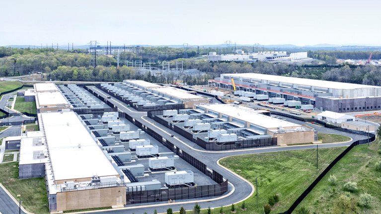 A sprawling data center complex surrounded by greenery and infrastructure. Several rectangular buildings with white roofs are visible, densely packed with cooling units or similar equipment, with at least one building under construction.