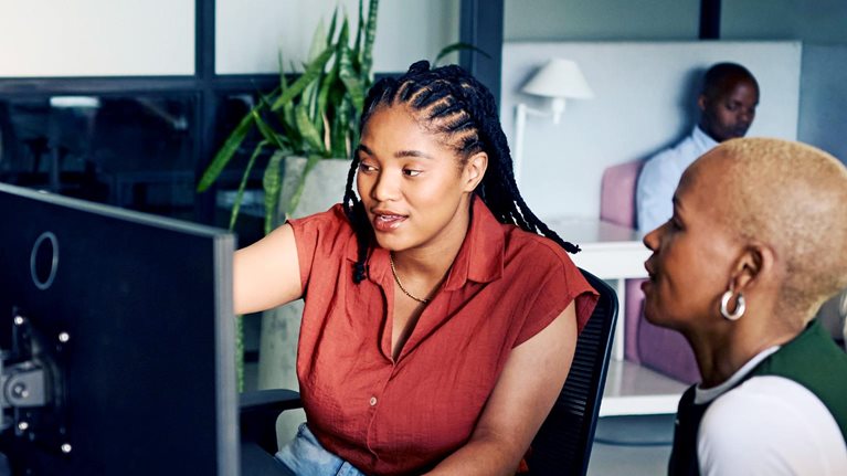 Two women collaborating closely at a computer, intensely focused on the screen. The one woman gestures towards the monitor, suggesting an learning or problem-solving session.