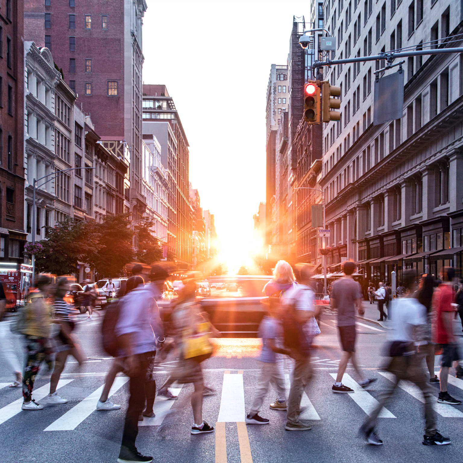 A diverse crowd of people walking through a bustling downtown intersection with the sunset in the background.