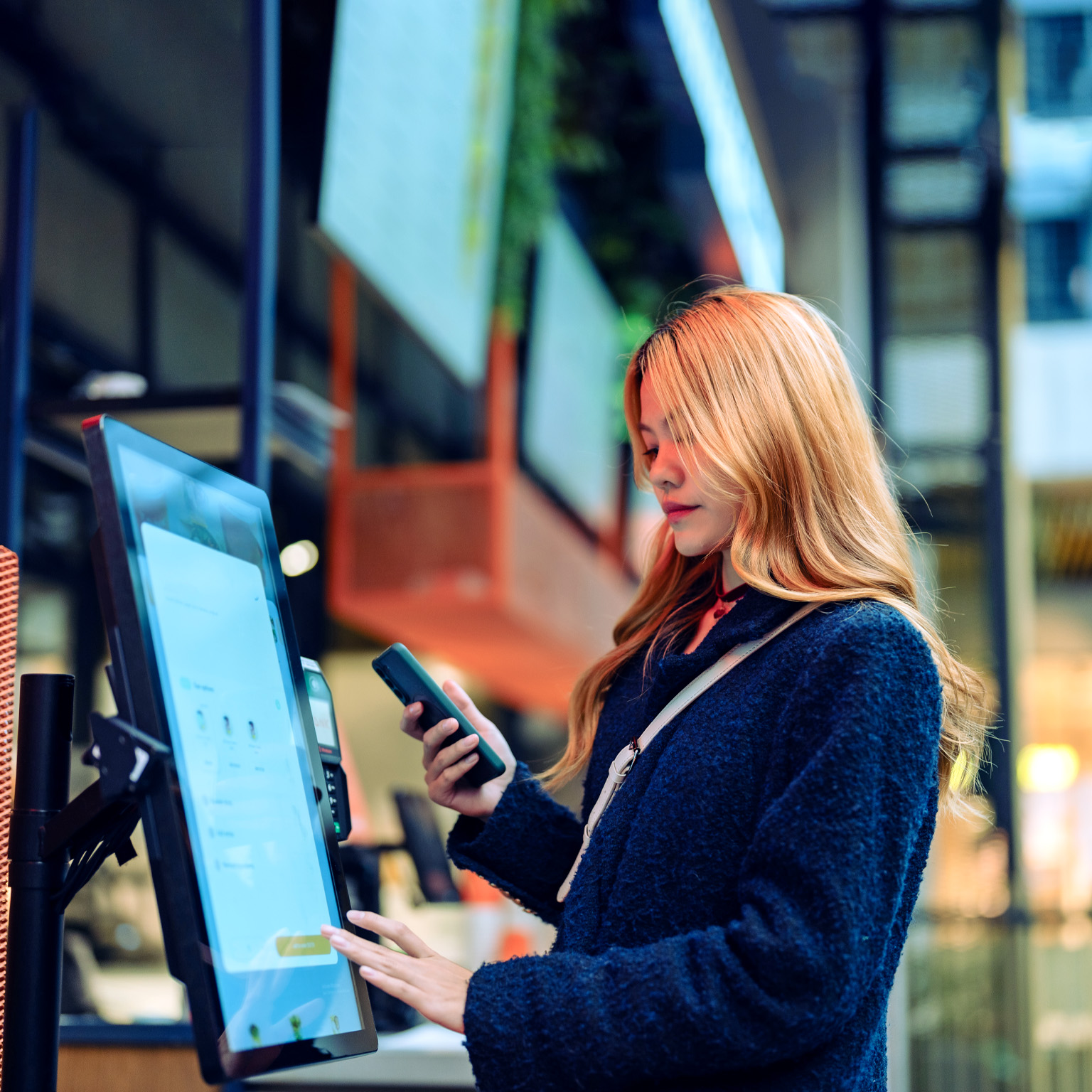 Women in casual clothing holding up her cell phone as she uses a smart checkout screen in a department store. 