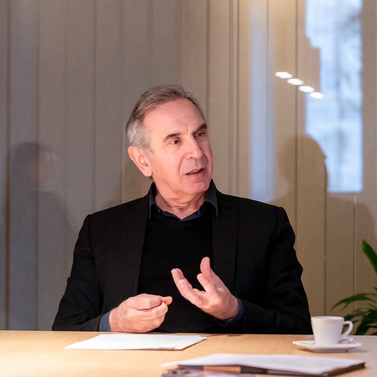 Carlo Capasa at a table, wearing a dark blazer and engaging in animated conversation, his hands gesturing expressively.