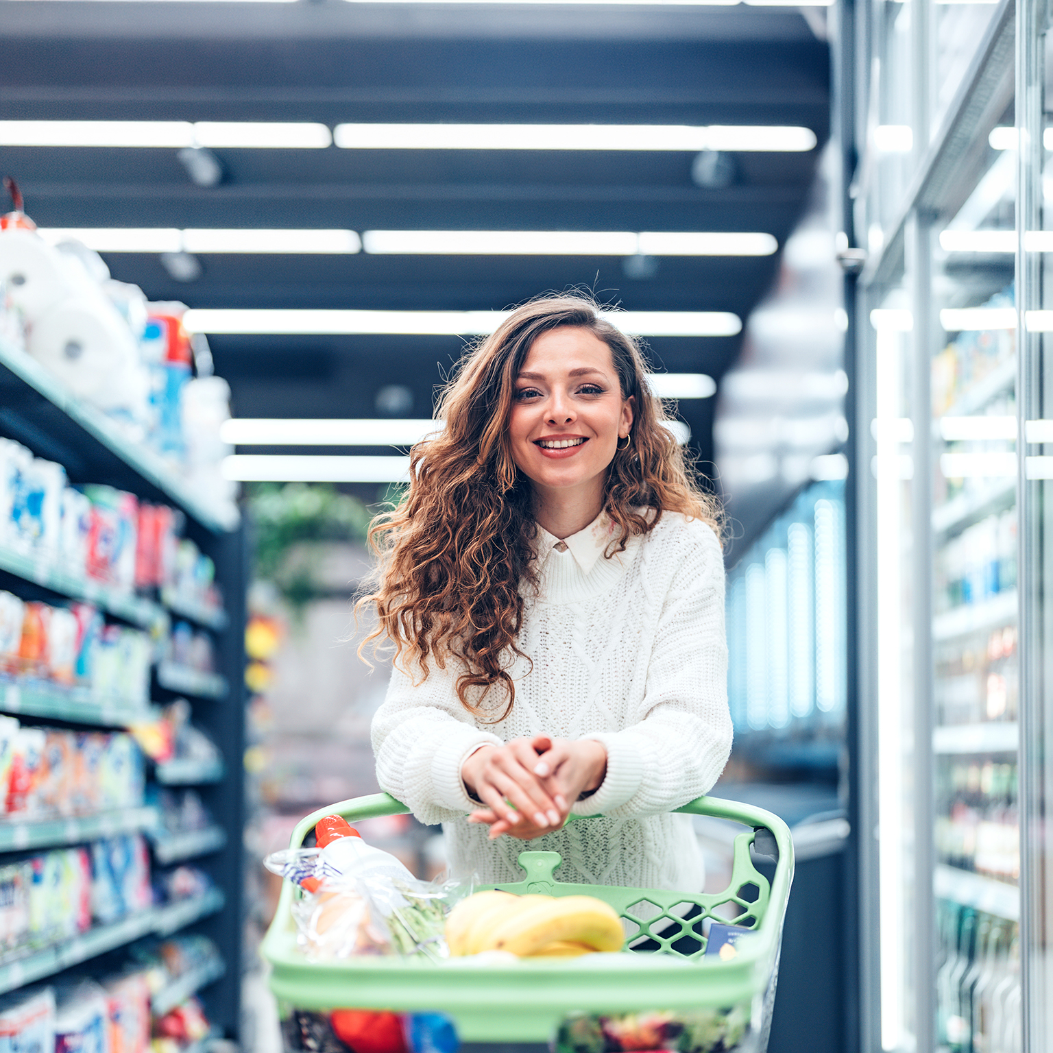A young woman stands in a bright, well-organized grocery store, carefully selecting products from the shelves. Surrounded by neatly arranged goods and fresh selections, the scene captures everyday consumer behavior, independence, and healthy decision-making in a contemporary retail environment.