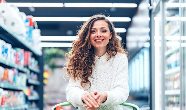 A young woman stands in a bright, well-organized grocery store, carefully selecting products from the shelves. Surrounded by neatly arranged goods and fresh selections, the scene captures everyday consumer behavior, independence, and healthy decision-making in a contemporary retail environment.