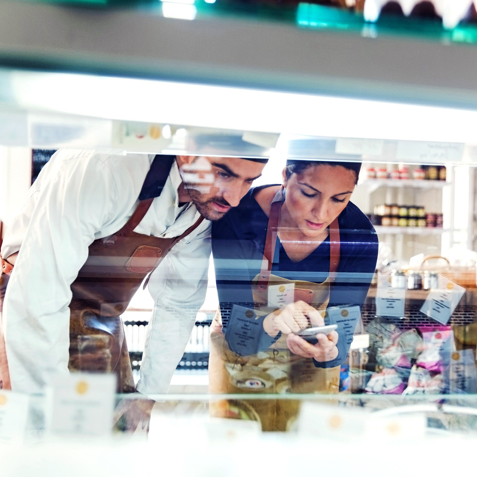 Male and female business owners wearing smocks using smart phone while examining display cabinet in a grocery store