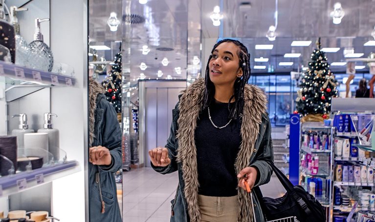 A woman walking down an aisle in a store while looking at personal care items. She is wearing a warm coat and holding a shopping basket, store holiday decorations can be seen in the background.