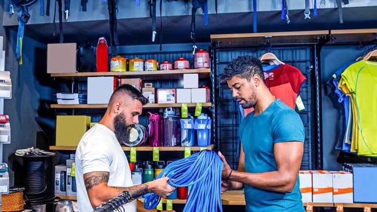 Two men examining a coil of blue climbing rope in an outdoor sports store, surrounded by shelves of gear and clothing. The shelves are stocked with various items, including water bottles, camping supplies, and colorful shirts.
