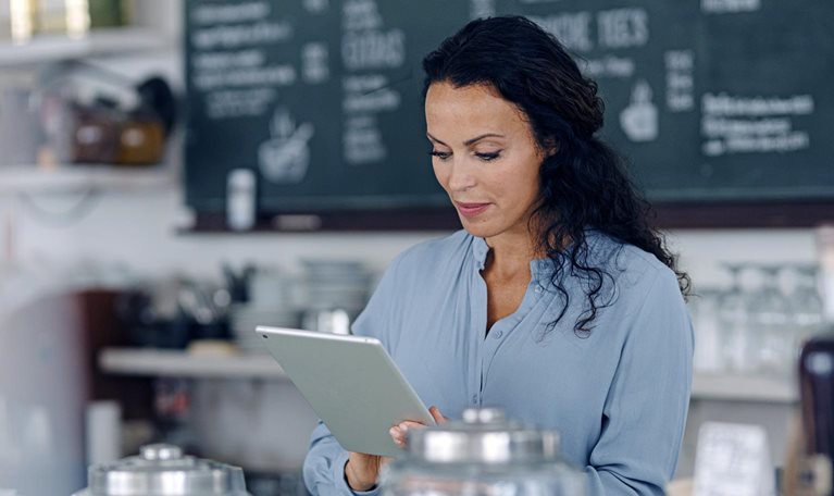 Brunette woman using digital tablet while standing at counter in coffee shop with the menu behind her.